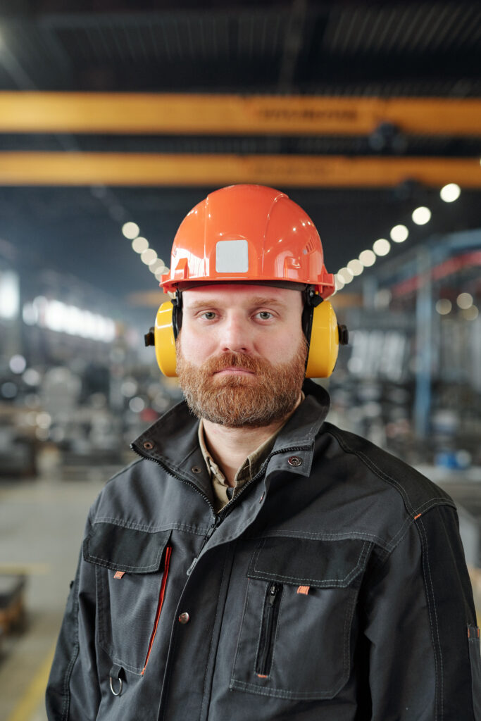 portrait-serious-young-bearded-man-work-jacket-protective-hardhat-standing-factory-shop